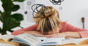 A person with their head resting on a desk covered in open books, with cartoon thought bubbles and face icons above their head.