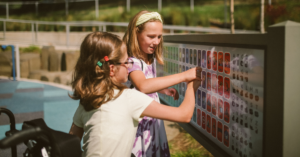 Two children interact with a large interactive display board featuring letters and numbers outdoors.
