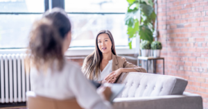 A woman in a tan jacket speaks to another person sitting across from her in a bright room with a brick wall and plants.