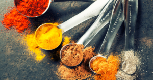 A collection of measuring spoons filled with various colorful spices, including red paprika, yellow turmeric, brown cinnamon, and white pepper, scattered on a dark surface.