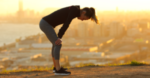 A person in athletic wear bends over with hands on knees, head down, during a sunrise or sunset with a city skyline in the background.