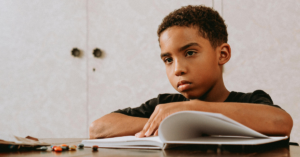 A child with curly hair wearing a black t-shirt rests their arms on an open book on a desk, with colorful pencils scattered nearby.