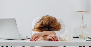 A person with reddish-brown hair rests their head on crossed arms on a white desk next to a laptop, phone, and crumpled tissues.