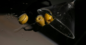 A martini glass tipped over with three olives on a dark, reflective surface with water droplets.