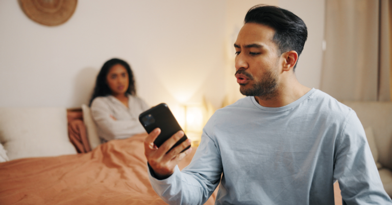 A man holds a smartphone in his hand and speaks to it with an angry expression, while a woman sits in the background on a bed with her arms crossed.