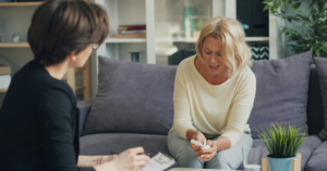 A person with blonde hair is sitting on a couch, holding tissues and appears to be upset while talking to someone off-camera who is holding a notepad.