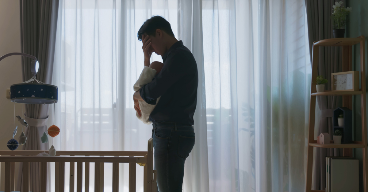 A man holding a baby and touching his forehead in frustration, standing next to a crib in front of a window with sheer curtains.